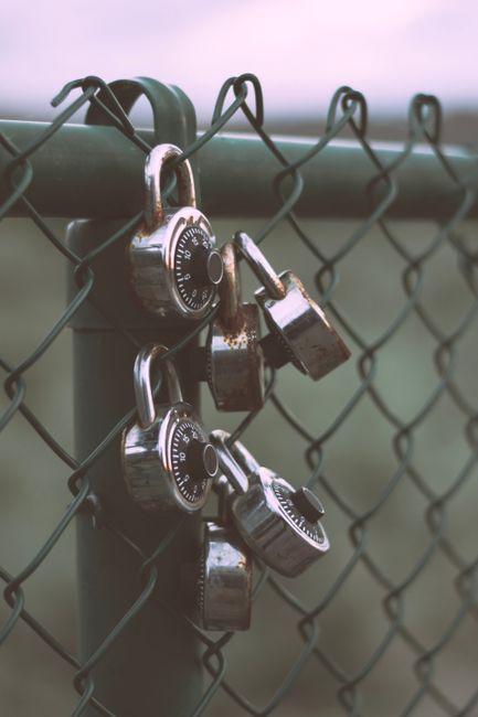 A close-up view of a metal chain-link fence with three combination padlocks attached to the top horizontal bar, each padlock featuring a circular dial with numbered increments, some with visible rust and signs of weathering. The locks are positioned in the foreground, with a blurred background of open landscape and soft, natural lighting that suggests an outdoor setting. The scene conveys a secure barrier, often associated with private property or restricted areas, aligning with themes of site access control in waste management or rubbish removal services by companies such as Waste Clearance Enfield. The image emphasizes the importance of controlled access for efficient, independent waste collection or on-site clearance activities, indirectly supporting the context of private disposal or alternative rubbish handling solutions. The overall tone remains neutral and professional, focusing on the hardware and environmental context without additional embellishment.