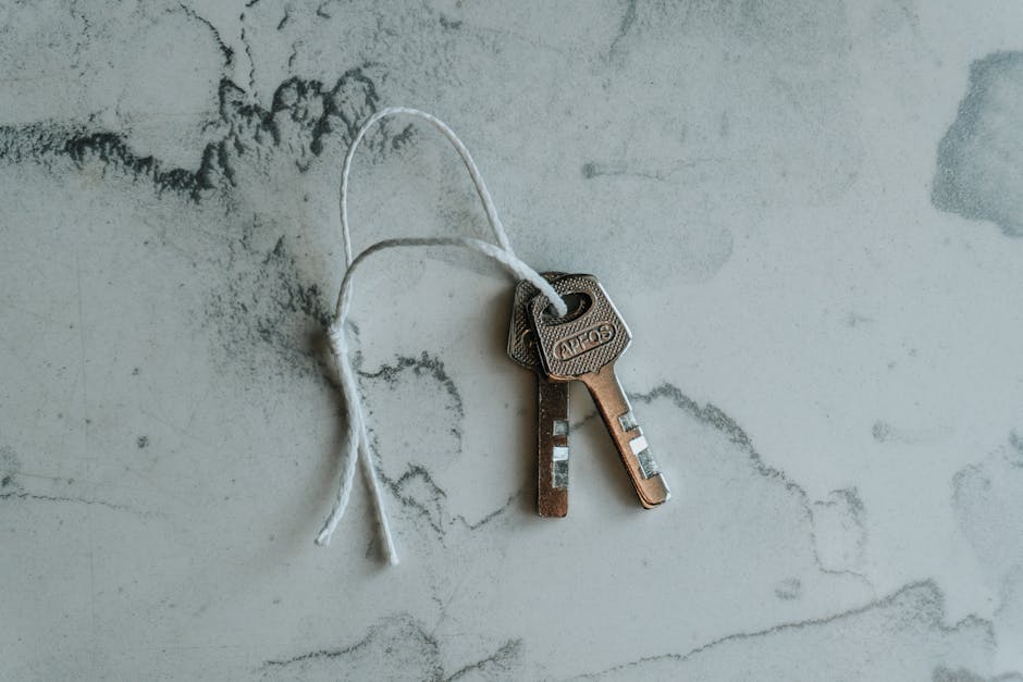 A close-up view of two identical brass keys with rectangular heads and a series of notches along the shanks, attached together with a loose white string. The keys are lying flat on a textured, light-colored surface that appears to be a marble or stone countertop with grey and white veining. The background is minimal, with no other objects visible, emphasizing the metallic finish and the simple, utilitarian design of the keys. The lighting is soft and natural, highlighting the metallic sheen of the brass and the subtle surface details. This image subtly relates to the theme of access or clearance, which aligns with the concept of rubbish removal and private waste handling services offered by Waste Clearance Enfield, providing an impression of control and access to secured areas.