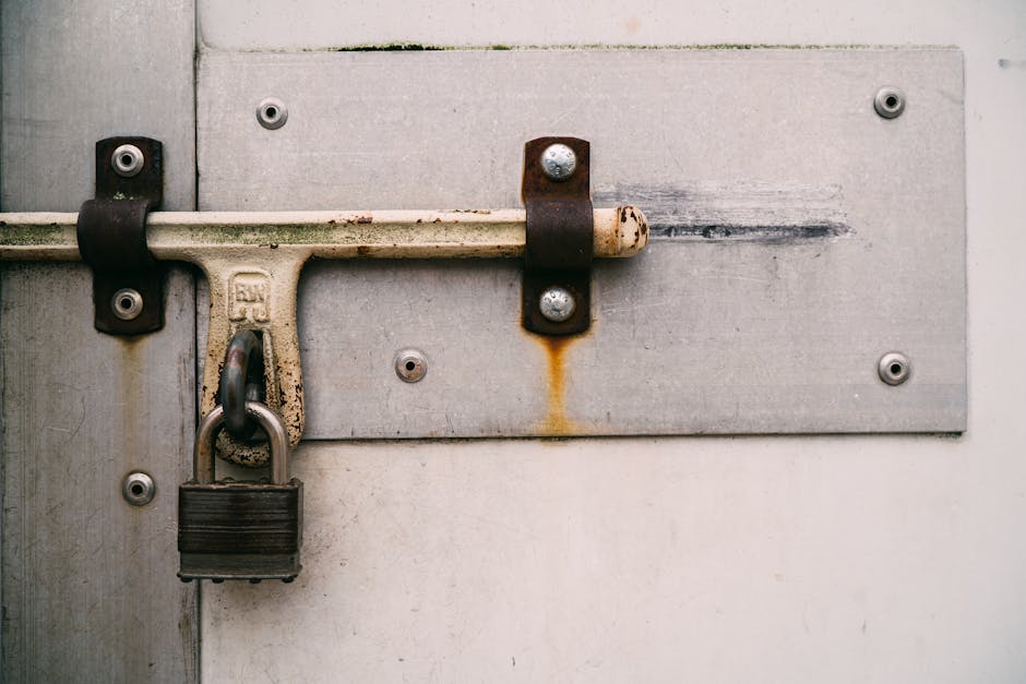 A close-up view of a metal chain-link fence with three combination padlocks attached to the top horizontal bar, each padlock featuring a circular dial with numbered increments, some with visible rust and signs of weathering. The locks are positioned in the foreground, with a blurred background of open landscape and soft, natural lighting that suggests an outdoor setting. The scene conveys a secure barrier, often associated with private property or restricted areas, aligning with themes of site access control in waste management or rubbish removal services by companies such as Waste Clearance Enfield. The image emphasizes the importance of controlled access for efficient, independent waste collection or on-site clearance activities, indirectly supporting the context of private disposal or alternative rubbish handling solutions. The overall tone remains neutral and professional, focusing on the hardware and environmental context without additional embellishment.