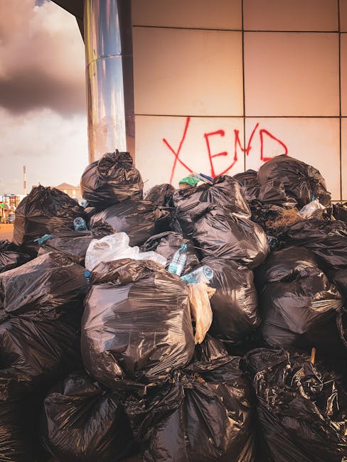 A large pile of black rubbish bags filled with household or garden waste, stacked along a modern building with tiled exterior walls. Some bags are partially torn, revealing plastic bottles and other debris inside. The bags are arranged on the pavement or possibly a driveway, with a few plastic water bottles scattered around the base. On the wall behind the bags, there is red graffiti reading 'XEND' spray-painted in large letters. The scene is illuminated by natural daylight, with a cloudy sky visible in the background, indicating outdoor urban surroundings. The image highlights issues of unmanaged waste accumulation that could relate to private rubbish collection or on-site clearance services such as those provided by Waste Clearance Enfield, supporting the context of alternative waste handling outside local authority collection in the Enfield area.