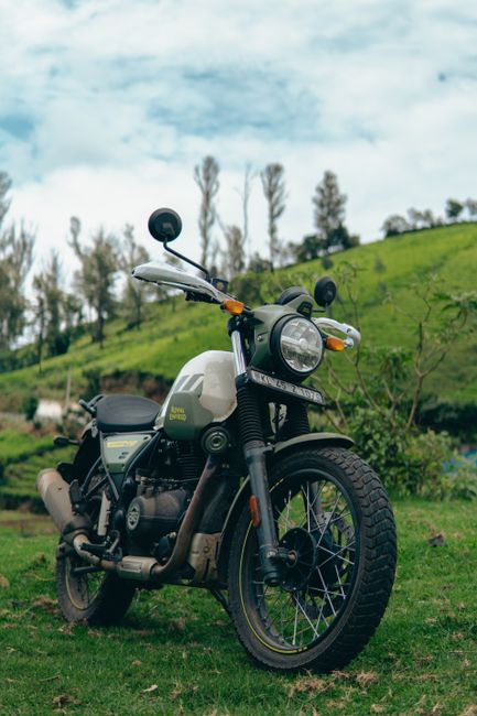 A black and white motorcycle with a matte finish is parked on a grassy area, facing slightly to the right. The motorcycle features a round headlight, black front forks, and spoked wheels with knobby tires suitable for rough terrain. The engine and exhaust system are visible with a dark finish, and the seat appears of black leather or synthetic material. In the background, there are green rolling hills with a few tall, thin trees and a partly cloudy, light blue sky. The scene is illuminated by natural daylight, highlighting the motorcycle's details and the vibrant greenery of the landscape. This setting suggests an environment suitable for private or on-site waste clearance activities, aligning with the context of independent rubbish removal services offered by Waste Clearance Enfield, emphasizing the possibility of off-road or rural waste collection options.