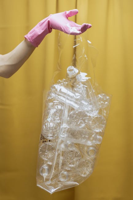 A person's hand, wearing a pink rubber glove, is seen holding a clear plastic rubbish bag filled with empty plastic bottles and containers. The plastic waste includes various bottles with caps and different shapes, some crushed or deformed, and is visibly transparent and glossy. The background consists of a plain, light-yellow curtain or wall, providing a neutral backdrop that highlights the plastic waste. The scene appears to take place indoors, possibly as part of a recycling or waste collection process, with the focus on segregating plastic refills for disposal or recycling. The person’s arm is extended, gripping the top of the bag securely, emphasizing the handling of waste materials in preparation for removal or recycling, aligning with the context of waste clearance or alternative rubbish removal services offered by Waste Clearance Enfield.