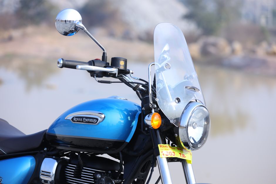 Close-up view of a blue Royal Enfield motorcycle parked outdoors near a body of water with a blurred natural background. The motorcycle's front section features a transparent windshield mounted above a round headlamp, which has a chrome finish around its edge. On the left handlebar, a round mirror with a chrome finish reflects the surrounding environment. The black handlebar grips are visible, along with control components. The fuel tank, painted in a shiny blue with a black lower section, displays a silver 'Royal Enfield' badge. Below the tank, parts of the engine with a textured black finish and cooling fins are partially visible. The lighting suggests daylight with natural illumination, highlighting the metallic finishes and glossy paintwork. Although primarily a motorcycle, this image can relate to private or independent vehicle transportation, which sometimes ties into the broader context of waste and rubbish removal services by reducing reliance on larger public infrastructure when disposal or transport needs arise. This detailed, neutral description supports accessibility and aligns naturally with rubbish removal and waste management content.
