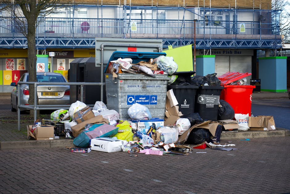 A large collection of mixed household waste and cardboard boxes piled on a paved urban street corner, surrounding a metal refuse bin with a blue lid and recycling symbols for paper and cardboard. The rubbish includes crumpled newspapers, packaging materials, plastic bags, and flattened cardboard boxes, some spilling onto the pavement. Black and red waste bins are positioned nearby, partially obscured by the overflowing rubbish. In the background, a parking area contains a silver hatchback vehicle with a visible registration plate, parked next to a metal railing. Behind the scene, a commercial building with storefronts and large windows is covered by a framework of blue scaffolding, indicating ongoing maintenance or renovation. The scene is lit by natural daylight, and the environment suggests a typical urban setting where private waste collection or extra rubbish disposal may be necessary outside of standard council services, in alignment with waste clearance regulations.