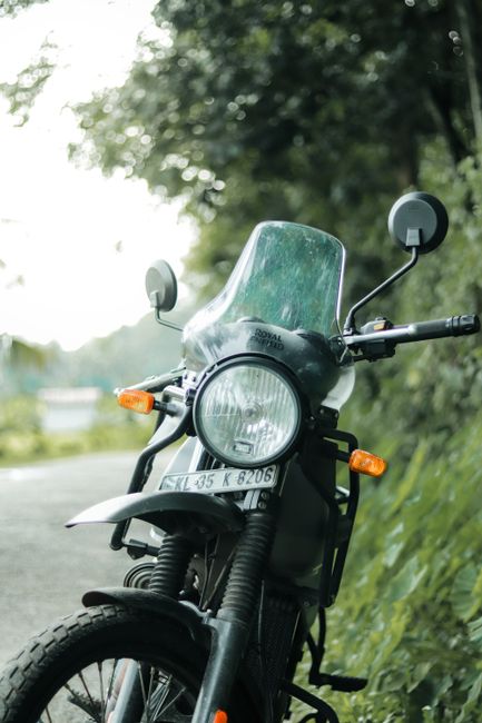 Close-up view of a blue Royal Enfield motorcycle parked outdoors near a body of water with a blurred natural background. The motorcycle's front section features a transparent windshield mounted above a round headlamp, which has a chrome finish around its edge. On the left handlebar, a round mirror with a chrome finish reflects the surrounding environment. The black handlebar grips are visible, along with control components. The fuel tank, painted in a shiny blue with a black lower section, displays a silver 'Royal Enfield' badge. Below the tank, parts of the engine with a textured black finish and cooling fins are partially visible. The lighting suggests daylight with natural illumination, highlighting the metallic finishes and glossy paintwork. Although primarily a motorcycle, this image can relate to private or independent vehicle transportation, which sometimes ties into the broader context of waste and rubbish removal services by reducing reliance on larger public infrastructure when disposal or transport needs arise. This detailed, neutral description supports accessibility and aligns naturally with rubbish removal and waste management content.