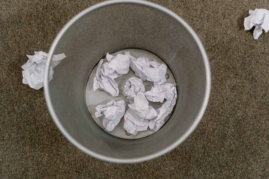 A cylindrical metal waste bin with a matte silver finish is shown from a top-down perspective, placed on a brown, textured carpeted floor. Inside the bin, several crumpled white paper sheets are loosely arranged, with some partially submerged in a small amount of water at the bottom. Two additional crumpled paper pieces are positioned outside the bin, resting on the carpet near the opening. The scene emphasizes a typical indoor office environment and highlights waste disposal practices related to rubbish collection services such as those offered by Waste Clearance Enfield, which provides alternative waste handling solutions for small businesses requiring prompt and efficient rubbish removal to maintain a tidy workspace.
