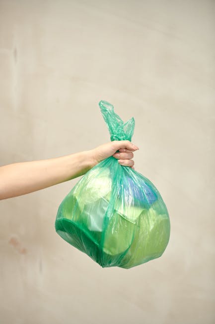 A person's arm extends from the left side of the image, holding a green and blue plastic rubbish bag by its twisted top. The bag appears to contain lightweight waste materials and is slightly translucent, showing some internal shapes and contents. The background is a plain, light beige wall, with no additional objects or environmental features visible. The scene is lit with soft, natural lighting, creating minimal shadows and emphasizing the bag's crinkled texture. This image relates to waste collection and rubbish removal services, such as those offered by Waste Clearance Enfield, highlighting the process of private rubbish disposal and alternative waste handling methods outside of municipal collection. The focus is solely on the bag and the hand, with no other objects or environmental context included, illustrating a typical scene of small-scale waste disposal for domestic or small business needs.